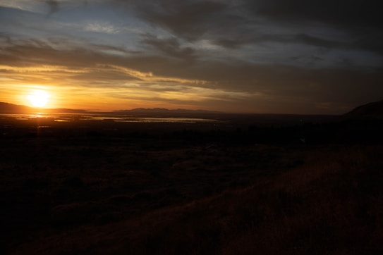 The landscape displays a breathtaking sunset over a vast, open terrain. The sun is setting near the horizon, casting a warm golden glow across the sky and reflecting on a distant body of water. The foreground is a darkened field or plain, creating a stark contrast with the vibrant colors of the sunset. Clouds are scattered, adding texture and depth to the sky.