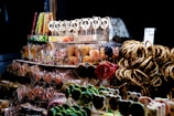 A display of assorted sweets, including gingerbread cookies decorated with icing, panda-shaped lollipops, licorice sticks, and pretzels. The items are arranged on shelves, with some wrapped in clear packaging. The scene has a festive and colorful atmosphere highlighted by a dark background.