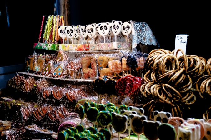 A display of assorted sweets, including gingerbread cookies decorated with icing, panda-shaped lollipops, licorice sticks, and pretzels. The items are arranged on shelves, with some wrapped in clear packaging. The scene has a festive and colorful atmosphere highlighted by a dark background.