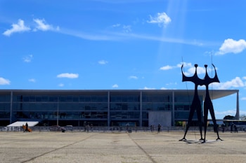 A modernist building with a facade of large glass windows is in the background. In front, a distinctive, abstract black sculpture stands tall under a clear blue sky with scattered white clouds. Several people walk around the open paved area in front of the building.