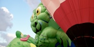 Large inflatable balloons depicting cartoon-like green creatures with playful expressions are prominently featured. A red and white hot air balloon can also be seen, set against a partially cloudy blue sky.