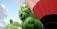 Large inflatable balloons depicting cartoon-like green creatures with playful expressions are prominently featured. A red and white hot air balloon can also be seen, set against a partially cloudy blue sky.