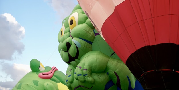 Large inflatable balloons depicting cartoon-like green creatures with playful expressions are prominently featured. A red and white hot air balloon can also be seen, set against a partially cloudy blue sky.