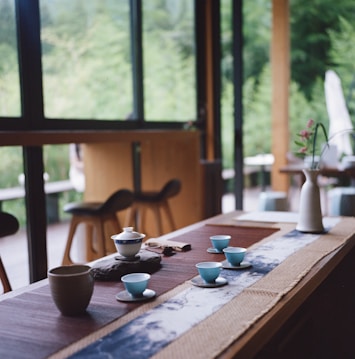 A serene tea setting on a wooden table with several small teacups and a teapot, accompanied by a vase with flowers. The table is adorned with a decorative runner featuring a blue and white pattern. Large windows in the background reveal a lush green garden, allowing natural light to softly illuminate the room.