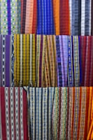 A collection of colorful woven fabrics is neatly arranged in rows. The fabrics feature various patterns, including stripes and geometric designs, with a vibrant range of colors including reds, blues, yellows, and purples. Each fabric has a unique pattern and color combination, creating a visually rich tapestry.