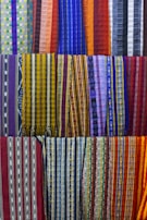 A collection of colorful woven fabrics is neatly arranged in rows. The fabrics feature various patterns, including stripes and geometric designs, with a vibrant range of colors including reds, blues, yellows, and purples. Each fabric has a unique pattern and color combination, creating a visually rich tapestry.