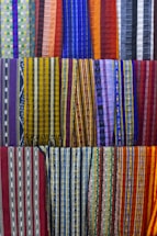 A collection of colorful woven fabrics is neatly arranged in rows. The fabrics feature various patterns, including stripes and geometric designs, with a vibrant range of colors including reds, blues, yellows, and purples. Each fabric has a unique pattern and color combination, creating a visually rich tapestry.