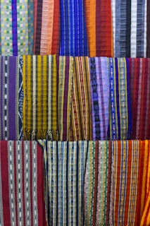 A collection of colorful woven fabrics is neatly arranged in rows. The fabrics feature various patterns, including stripes and geometric designs, with a vibrant range of colors including reds, blues, yellows, and purples. Each fabric has a unique pattern and color combination, creating a visually rich tapestry.