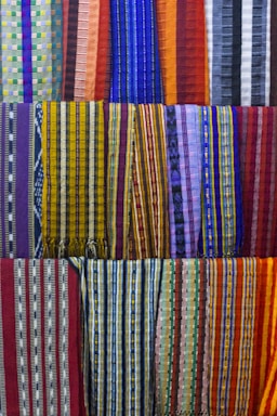 A collection of colorful woven fabrics is neatly arranged in rows. The fabrics feature various patterns, including stripes and geometric designs, with a vibrant range of colors including reds, blues, yellows, and purples. Each fabric has a unique pattern and color combination, creating a visually rich tapestry.