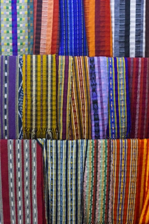 A collection of colorful woven fabrics is neatly arranged in rows. The fabrics feature various patterns, including stripes and geometric designs, with a vibrant range of colors including reds, blues, yellows, and purples. Each fabric has a unique pattern and color combination, creating a visually rich tapestry.