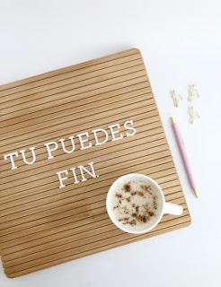 A wooden letter board with the words 'TU PUEDES FIN.' in white letters. A cup of frothy coffee sits on the board with cinnamon sprinkled on top. On the right, there is a pink pen with gold accents and three gold paper clip flamingos on a white background.