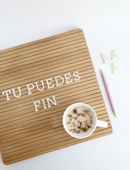 A wooden letter board with the words 'TU PUEDES FIN.' in white letters. A cup of frothy coffee sits on the board with cinnamon sprinkled on top. On the right, there is a pink pen with gold accents and three gold paper clip flamingos on a white background.