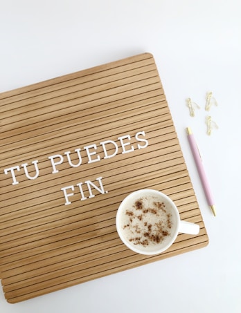A wooden letter board with the words 'TU PUEDES FIN.' in white letters. A cup of frothy coffee sits on the board with cinnamon sprinkled on top. On the right, there is a pink pen with gold accents and three gold paper clip flamingos on a white background.