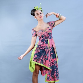A woman poses in a colorful, patterned dress with intricate floral designs and a unique asymmetrical cut, featuring pink and black colors with green accents. Her hair is styled in an elegant updo adorned with a green ribbon, and she accessorizes with bright bracelets on her wrist. The background is a simple light blue, drawing focus to the vibrant outfit.