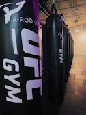 A row of black punching bags hangs in a gym, with clear branding on the front. The gym has an industrial vibe with exposed ceilings and a mix of artificial lighting adding purplish and yellowish hues.