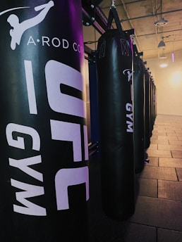 A row of black punching bags hangs in a gym, with clear branding on the front. The gym has an industrial vibe with exposed ceilings and a mix of artificial lighting adding purplish and yellowish hues.