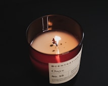 A lit candle with a visible flame sits in a dark glass container. The label on the container reads 'Scentationals' and 'Onyx No. 15' and mentions natural wax with a description of the fragrance. The candle is placed on a dark surface, creating a contrast with the light from the flame.
