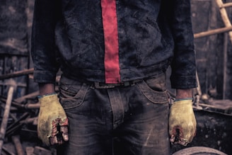A person wearing dirty work clothes stands with their hands visible. The individual is wearing a dark jacket with a red stripe down the center, and yellow gloves covering their hands. The surroundings appear to be cluttered with various tools and materials in a workshop-like environment.