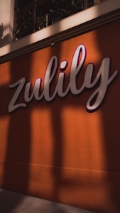 A large sign with the word 'Zulily' is displayed prominently against an orange wall. The sign features cursive white lettering with a pink outline. Shadows cast by nearby objects create an interesting pattern on the wall.
