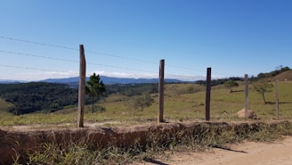 A rural landscape featuring a barbed wire fence constructed with wooden posts. Rolling hills stretch into the distance with patches of green grass and scattered trees under a clear blue sky.