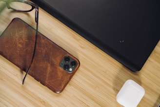 A wooden surface is partially covered by a leather-cased smartphone, a pair of eyeglasses resting on the phone, a corner of a black laptop, and a pair of wireless earbuds in a charging case. The arrangement suggests a neat workspace or personal tech collection.