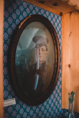 A framed, vintage portrait of a military officer in uniform hangs on a floral-patterned wallpaper. The framing is oval, with a dark, ornate wooden border. To the side, there is a small, partially visible name plaque attributing the photo. A glass vase with a single green leaf rests on a wooden shelf nearby, contributing to the rustic setting.