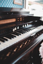 An antique piano with a detailed, dark wood finish is positioned indoors. The keyboard features evenly spaced black and white keys, while the upper panel includes decorative elements and several knobs with labels. A framed picture is visible on the patterned wallpaper background, adding a vintage vibe to the setting.