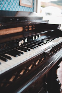 An antique piano with a detailed, dark wood finish is positioned indoors. The keyboard features evenly spaced black and white keys, while the upper panel includes decorative elements and several knobs with labels. A framed picture is visible on the patterned wallpaper background, adding a vintage vibe to the setting.