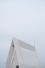 A minimalist, modern white building with a triangular roof and clean lines is set against a pale, overcast sky, featuring a small hanging light fixture and an antenna.