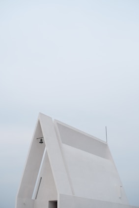 A minimalist, modern white building with a triangular roof and clean lines is set against a pale, overcast sky, featuring a small hanging light fixture and an antenna.
