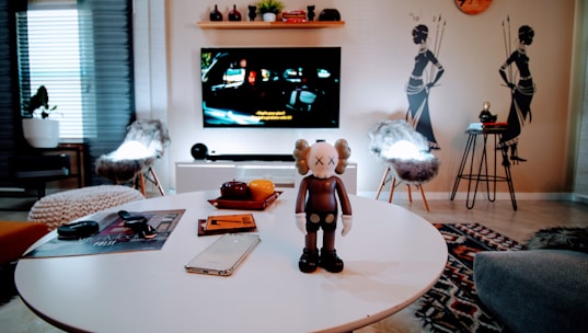 A modern living room with a round white table in the foreground, holding a brown and white cartoon-like figure with crossed-out eyes. Nearby are a smartphone and a notebook. The background features a television on a stand displaying a movie scene, while two chairs with furry covers are placed on either side. The wall behind has tribal-themed decals and a small shelf with decorative items.