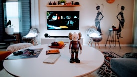 A modern living room with a round white table in the foreground, holding a brown and white cartoon-like figure with crossed-out eyes. Nearby are a smartphone and a notebook. The background features a television on a stand displaying a movie scene, while two chairs with furry covers are placed on either side. The wall behind has tribal-themed decals and a small shelf with decorative items.