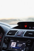 The image captures the interior of a car focused on the dashboard. A touchscreen display is playing music from an iPod, showing a song titled 'Little League' by Conan Gray from the album 'Kid Krow'. The dashboard includes various controls, vents, and a prominent hazard button.