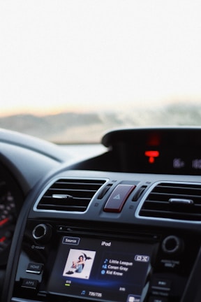 The image captures the interior of a car focused on the dashboard. A touchscreen display is playing music from an iPod, showing a song titled 'Little League' by Conan Gray from the album 'Kid Krow'. The dashboard includes various controls, vents, and a prominent hazard button.