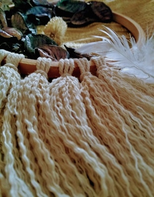 A close-up view of a textured textile or yarn intertwined with a hoop, adorned with dried leaves and a single white feather. The color palette is dominated by warm and earthy tones, enhancing the natural, rustic feel.