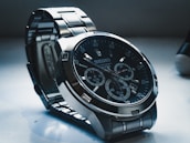 A detailed view of a Seiko wristwatch with a dark dial and metallic strap. The watch face includes multiple sub-dials with intricate markings and a date display. The lighting casts shadows, highlighting the watch's polished metal surfaces.