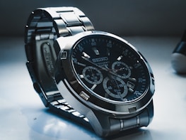 A detailed view of a Seiko wristwatch with a dark dial and metallic strap. The watch face includes multiple sub-dials with intricate markings and a date display. The lighting casts shadows, highlighting the watch's polished metal surfaces.