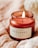 A burning scented candle labeled 'Bergamot' in a jar placed on a soft, beige fabric surface. The candle is reddish-brown in color and emits a gentle flame.