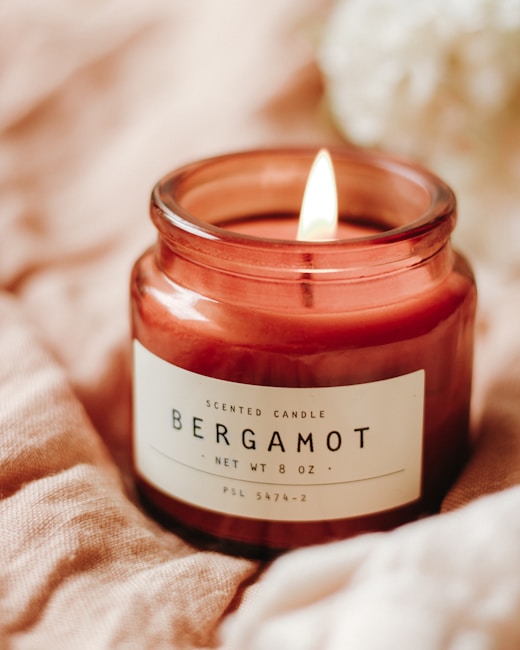 A burning scented candle labeled 'Bergamot' in a jar placed on a soft, beige fabric surface. The candle is reddish-brown in color and emits a gentle flame.