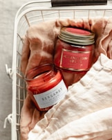 Two scented candles in jars labeled 'Bergamot' and 'Cinnamon Spice' are placed in a white wire basket lined with soft, beige fabric. The candles have a red hue, and the labels are prominently displayed.