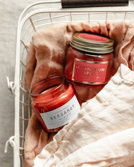 Two scented candles in jars labeled 'Bergamot' and 'Cinnamon Spice' are placed in a white wire basket lined with soft, beige fabric. The candles have a red hue, and the labels are prominently displayed.