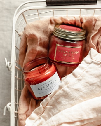 Two scented candles in jars labeled 'Bergamot' and 'Cinnamon Spice' are placed in a white wire basket lined with soft, beige fabric. The candles have a red hue, and the labels are prominently displayed.