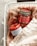 Two scented candles in jars labeled 'Bergamot' and 'Cinnamon Spice' are placed in a white wire basket lined with soft, beige fabric. The candles have a red hue, and the labels are prominently displayed.