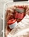 Two scented candles in jars labeled 'Bergamot' and 'Cinnamon Spice' are placed in a white wire basket lined with soft, beige fabric. The candles have a red hue, and the labels are prominently displayed.