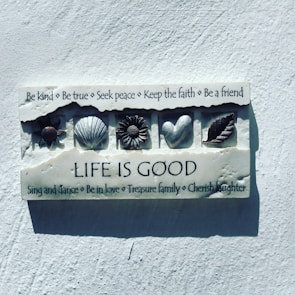 A decorative plaque on a wall contains various positive affirmations and symbols. The text encourages kindness, truth, peace, faith, and friendship. Below are icons of a turtle, shell, flower, heart, and leaf, followed by the phrase 'Life is Good'. Additional phrases promote dancing, love, family, and laughter.