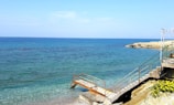 A serene seascape with clear blue waters stretching to the horizon. A stone staircase leads down to a rusty metal platform that extends into the sea. In the foreground, there are yellow flowers growing along the rocky shore. The sky is mostly clear with a few scattered clouds.