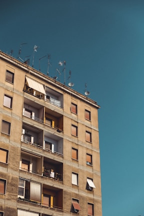 A beige residential building with multiple balconies and closed shutters is topped with various antennas under a clear blue sky.
