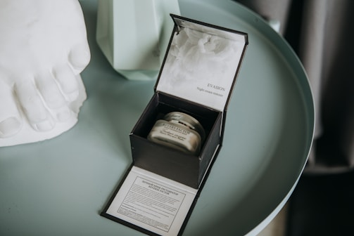 An elegant round table holds an open box with a cosmetic cream jar inside. Beside the box, there is a decorative white object resembling toes and a geometric vase with minimalist design.