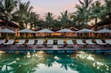 A serene resort setting features a series of lounge chairs lined in a row beside a reflective swimming pool. Large umbrellas provide shade, set against a backdrop of tall palm trees and a warm, glowing structure with expansive windows. The early evening sky casts a soft, calming glow over the entire scene.