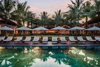 A serene resort setting features a series of lounge chairs lined in a row beside a reflective swimming pool. Large umbrellas provide shade, set against a backdrop of tall palm trees and a warm, glowing structure with expansive windows. The early evening sky casts a soft, calming glow over the entire scene.